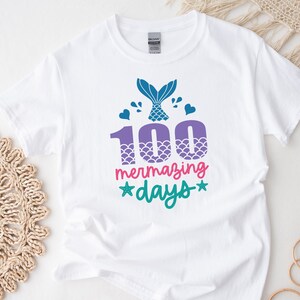 May include: White t-shirt featuring a mermaid tail graphic and the text "100 mermazing days" in pink, green, and purple.