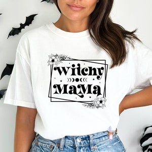 May include: White t-shirt with black text that reads "Witchy Mama" in a retro font. The text is framed by a black square with floral accents.
