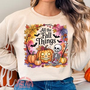 Puede incluir: Sudadera beige con un diseño colorido de Halloween. El gráfico incluye calabazas, un esqueleto, murciélagos y el texto "All the Fall Things" rodeado de hojas de otoño y telarañas.