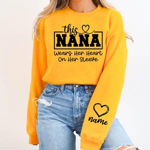 May include: A bright yellow sweatshirt with the text "this NANA wears her heart on her sleeve" in black script. A black heart and the word "name" are on the sleeve. The sweatshirt is paired with blue jeans.