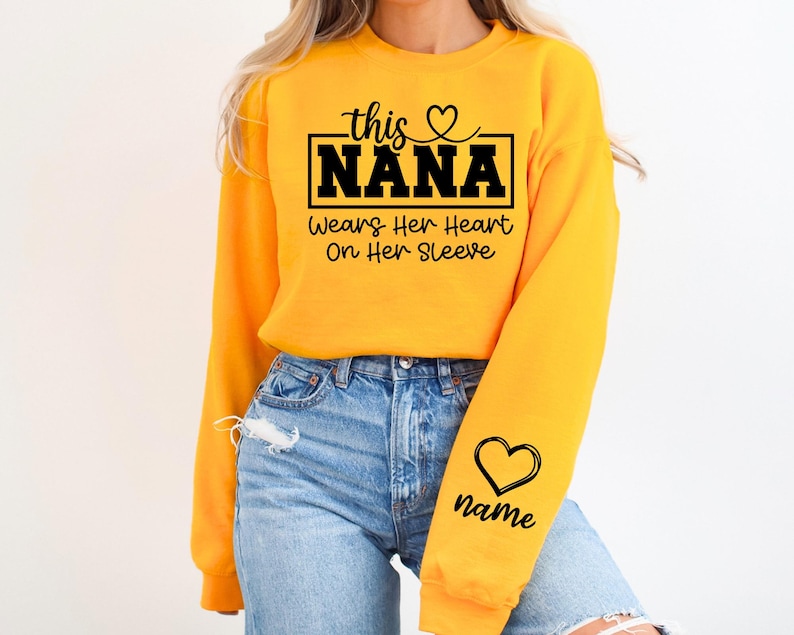 May include: A bright yellow sweatshirt with the text "this NANA wears her heart on her sleeve" in black script. A black heart and the word "name" are on the sleeve. The sweatshirt is paired with blue jeans.