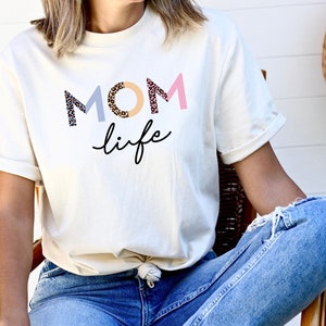 May include: A white t-shirt with the text "MOM life" in colorful letters with a leopard print design. The letters are in pink, blue, and beige.