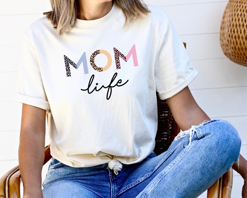 May include: A white t-shirt with the text "MOM life" in colorful letters with a leopard print design. The letters are in pink, blue, and beige.