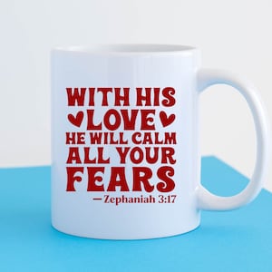 K&ouml;nnte beinhalten: Wei&szlig;e Keramiktasse mit einer roten Aufschrift, die lautet: "With His Love He Will Calm All Your Fears - Zephaniah 3:17".