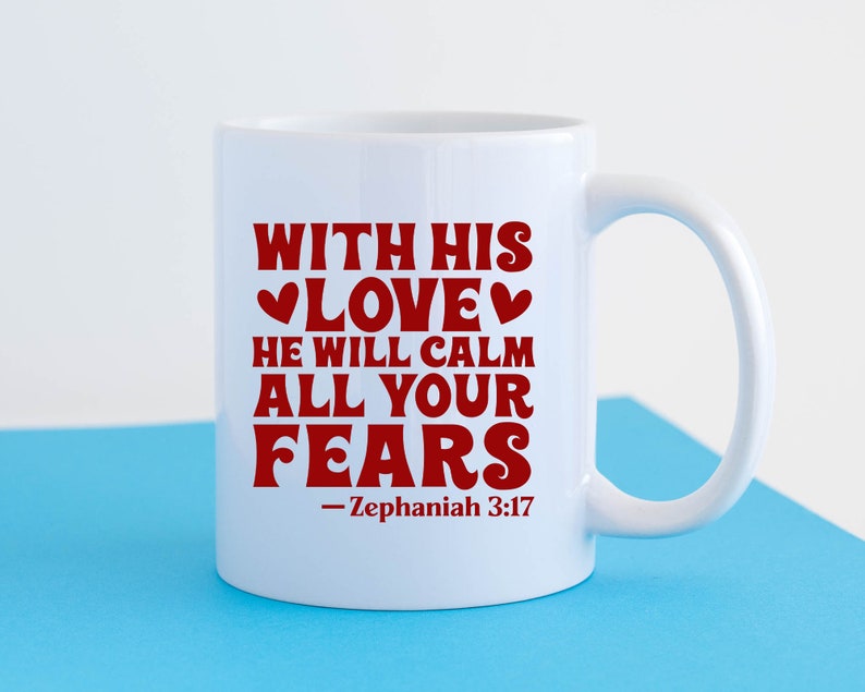 K&ouml;nnte beinhalten: Wei&szlig;e Keramiktasse mit einer roten Aufschrift, die lautet: "With His Love He Will Calm All Your Fears - Zephaniah 3:17".