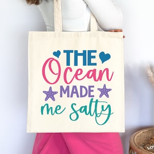 May include: A white tote bag with the text "The Ocean Made Me Salty" in pink, blue, purple, and teal lettering. The text is surrounded by two pink hearts, two blue hearts, and two purple starfish.