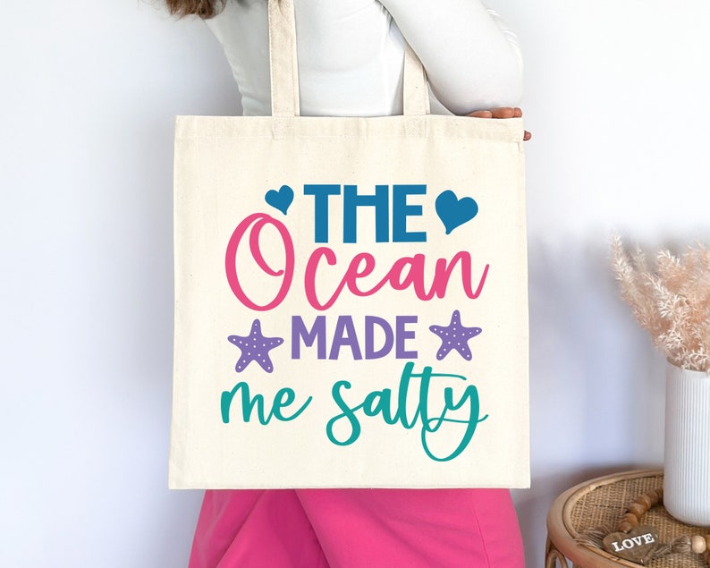 May include: A white tote bag with the text "The Ocean Made Me Salty" in pink, blue, purple, and teal lettering. The text is surrounded by two pink hearts, two blue hearts, and two purple starfish.