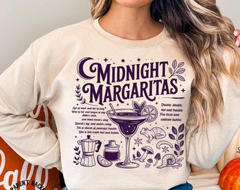 May include: A cream-coloured sweatshirt featuring a purple graphic with the words "Midnight Margaritas." The design includes a cocktail, a frog, mushrooms, and botanical elements. The text also includes a recipe. A fun, whimsical design.