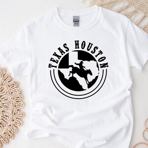 Op de afbeelding: Wit T-shirt met een zwarte afbeelding van een cowboy die op een paard rijdt voor de staat Texas. De afbeelding is omgeven door een cirkel met de woorden "Texas" en "Houston".