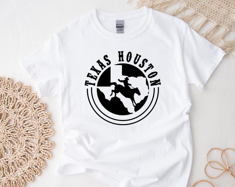 Op de afbeelding: Wit T-shirt met een zwarte afbeelding van een cowboy die op een paard rijdt voor de staat Texas. De afbeelding is omgeven door een cirkel met de woorden "Texas" en "Houston".