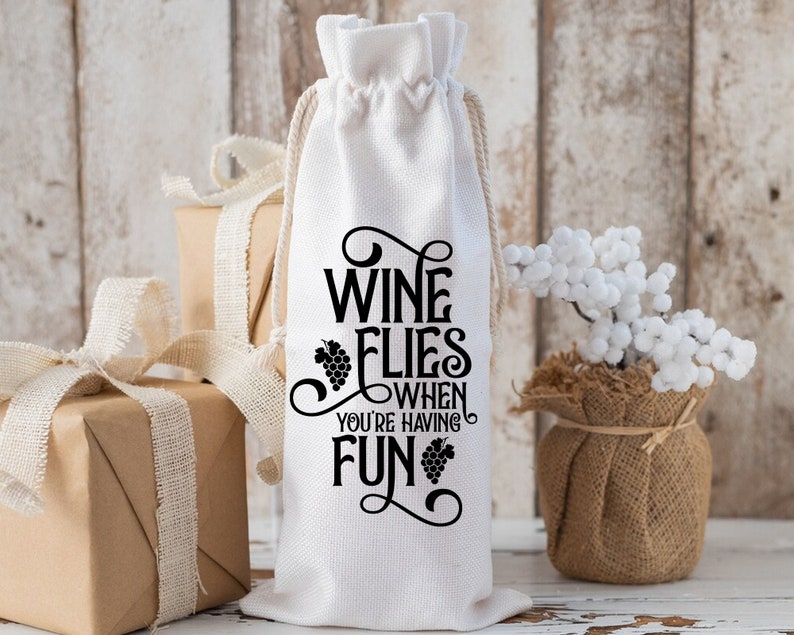 May include: White fabric wine bottle bag with black text that reads "Wine Flies When You're Having Fun" with a grape design.