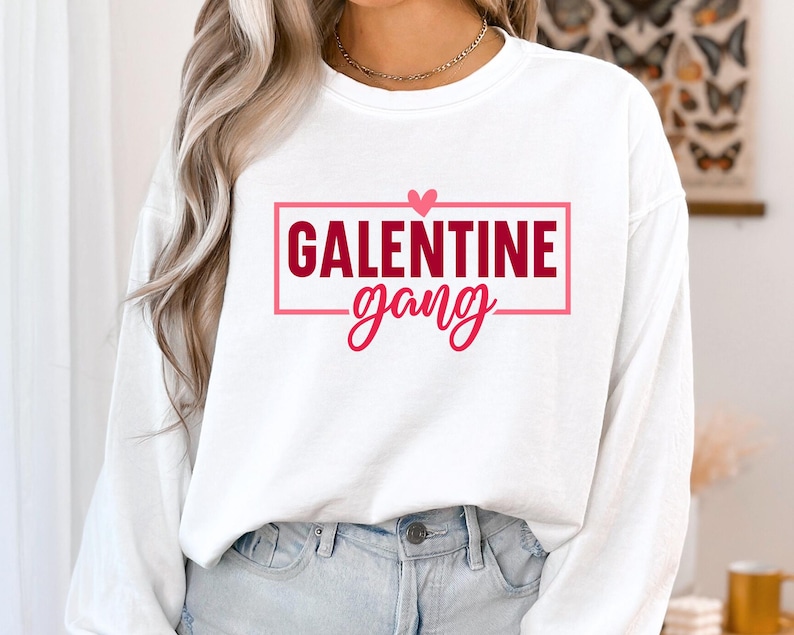 Op de afbeelding: Witte longsleeve trui met een rode doos en een roze hartgrafiek met de tekst "Galentine gang".