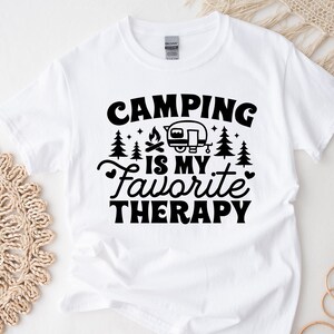 May include: White t-shirt with black text that reads "Camping is my favorite therapy". The text is surrounded by small images of trees and a campfire. A small black and white image of a camper is above the text.