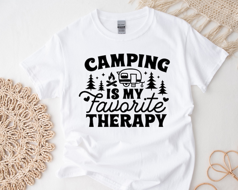 May include: White t-shirt with black text that reads "Camping is my favorite therapy". The text is surrounded by small images of trees and a campfire. A small black and white image of a camper is above the text.