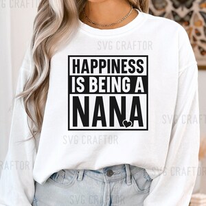 May include: White long-sleeve shirt with black text that reads "Happiness is being a Nana" with a small black heart. The shirt is worn by a person with long blonde hair.