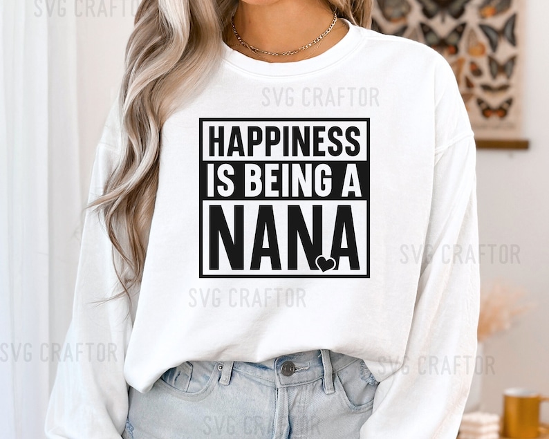 May include: White long-sleeve shirt with black text that reads "Happiness is being a Nana" with a small black heart. The shirt is worn by a person with long blonde hair.