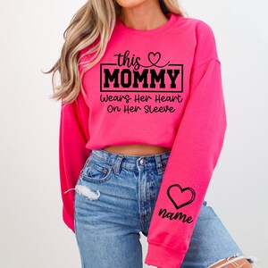 May include: A bright pink sweatshirt with the text "this MOMMY Wears Her Heart On Her Sleeve" in black. A black heart and the word "name" are printed on the sleeve. The sweatshirt is a crew neck style.