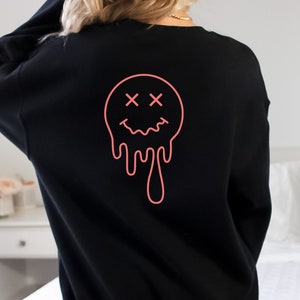 K&ouml;nnte beinhalten: Schwarzer Pullover mit einem pinkfarbenen, tropfenden Smiley-Grafik. Der Smiley hat X f&uuml;r Augen.