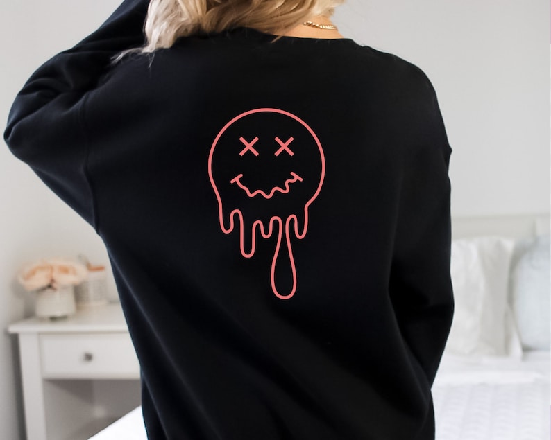 K&ouml;nnte beinhalten: Schwarzer Pullover mit einem pinkfarbenen, tropfenden Smiley-Grafik. Der Smiley hat X f&uuml;r Augen.