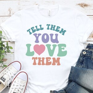 May include: White t-shirt with the text "Tell them you love them" in colorful retro font. The text is in shades of teal, purple, green, and orange. A pink heart is in the middle of the text.