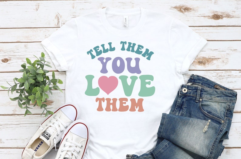 May include: White t-shirt with the text "Tell them you love them" in colorful retro font. The text is in shades of teal, purple, green, and orange. A pink heart is in the middle of the text.