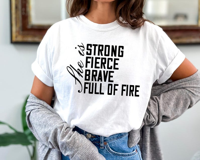 May include: White t-shirt with black text that reads "She is strong fierce brave full of fire".