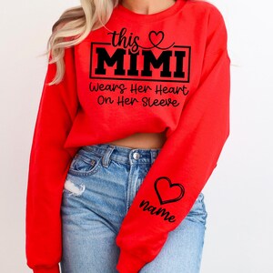 May include: A red sweatshirt with the text "this MIMI wears her heart on her sleeve" in black. A black heart and the word "name" are on the sleeve. The sweatshirt is worn with blue jeans.