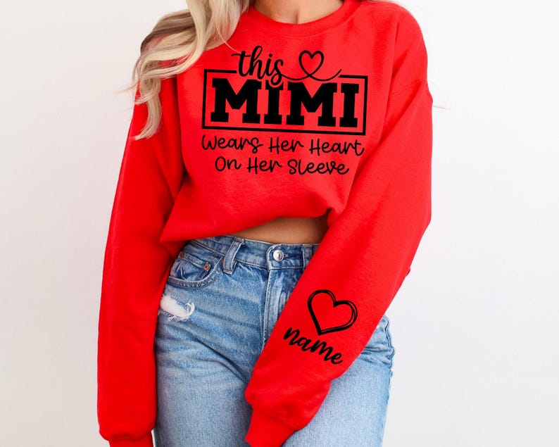 May include: A red sweatshirt with the text "this MIMI wears her heart on her sleeve" in black. A black heart and the word "name" are on the sleeve. The sweatshirt is worn with blue jeans.