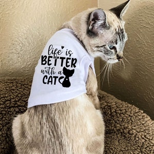 Pu&ograve; includere: Un gatto bianco che indossa una maglietta bianca con la scritta "life is better with a cat" e una silhouette nera di un gatto. Il gatto &egrave; seduto su una superficie di pelo marrone.