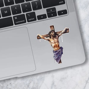 May include: A sticker of a man on a wooden cross. The man is depicted with a crown of thorns and blood on his body. The sticker is on a silver laptop.
