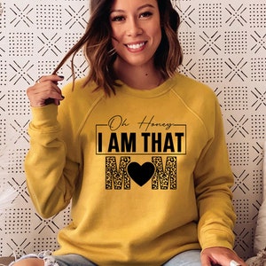 May include: A mustard yellow sweatshirt with a black graphic that says "Oh Honey I AM THAT MOM" with a black heart in the middle of the word MOM. The graphic is surrounded by a leopard print pattern.