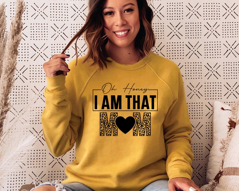 May include: A mustard yellow sweatshirt with a black graphic that says "Oh Honey I AM THAT MOM" with a black heart in the middle of the word MOM. The graphic is surrounded by a leopard print pattern.