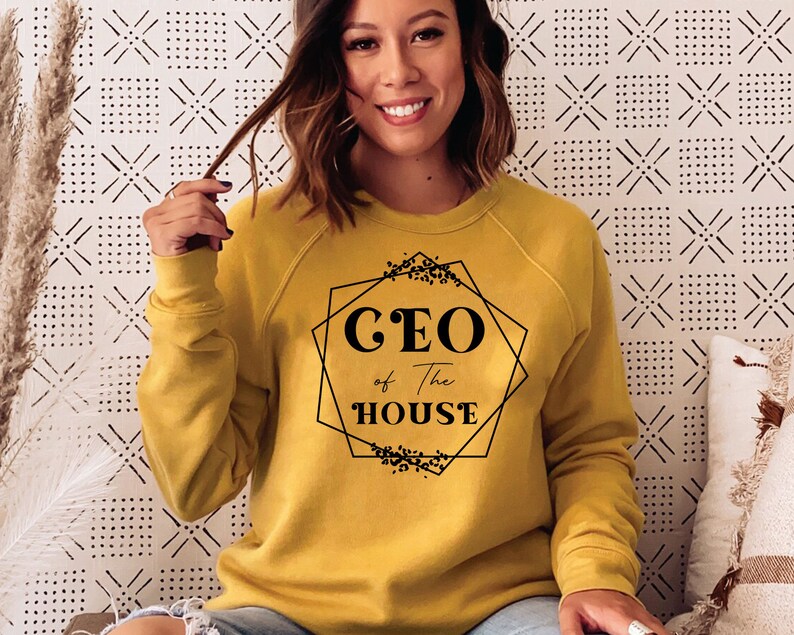 May include: A mustard yellow sweatshirt with black text that reads "CEO of the HOUSE" inside a geometric frame with floral accents.