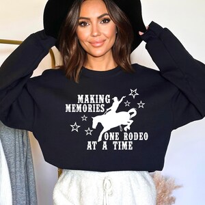 Op de afbeelding: Zwart gecropped sweatshirt met witte tekst die "Making Memories One Rodeo At A Time" luidt. In het midden van de tekst staat een wit silhouet van een persoon die een paard berijdt.