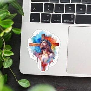 May include: A watercolor painting of Jesus Christ on the cross. The painting is in shades of blue, red, and orange, with splashes of white paint. Jesus is wearing a crown of thorns and has a pained expression on his face.