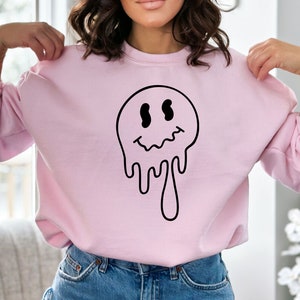 K&ouml;nnte beinhalten: Ein rosa Sweatshirt mit einem schwarzen, tropfenden Smiley-Motiv.