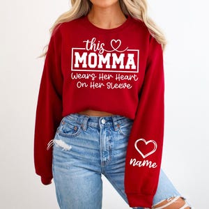 May include: A red sweatshirt with white text that reads "this MOMMA wears her heart on her sleeve." A white heart and the word "name" are on the right sleeve. The sweatshirt is paired with blue jeans.