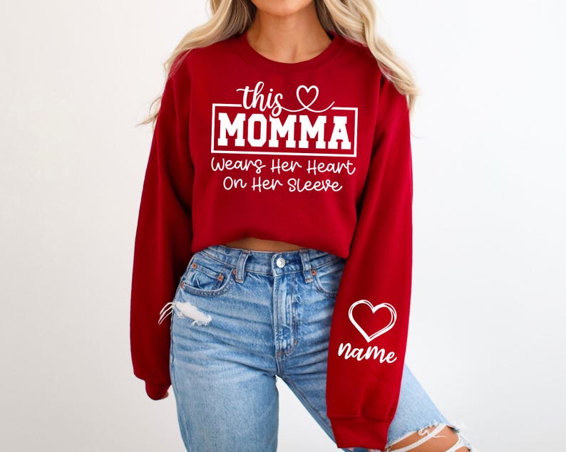 May include: A red sweatshirt with white text that reads "this MOMMA wears her heart on her sleeve." A white heart and the word "name" are on the right sleeve. The sweatshirt is paired with blue jeans.