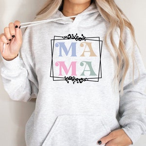 May include: A gray hoodie with a black frame and the word "MAMA" in pastel colors. The letters are in a retro font style and are surrounded by a floral design.