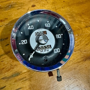 Puede incluir: Un velocímetro redondo Smiths con bisel cromado, esfera negra y números blancos del 20 al 100 MPH. El centro presenta un disco plateado, una aguja y un odómetro mecánico que muestra 50035. El texto incluye SMITHS y la palabra IGNITION.