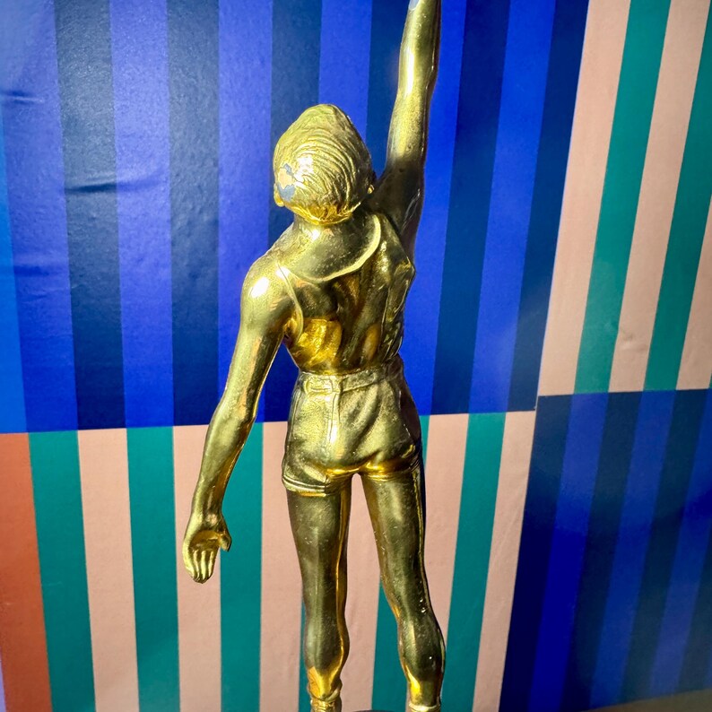 Puede incluir: Trofeo dorado que representa a una persona con pantalones cortos y camiseta sin mangas con un brazo levantado. El fondo presenta rayas azules, turquesas y rosas. Un s&iacute;mbolo de logro.