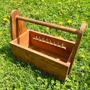 May include: A wooden tool caddy with a handle, set on a grassy surface. The caddy is rectangular with a series of small pegs inside. The wood has a natural finish. The handle is a rounded dowel. The caddy is designed to hold tools.
