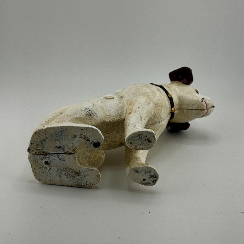Op de afbeelding: Een cr&egrave;me-kleurige gietijzeren bulldog-beeldje met een zwarte halsband en donker bordeauxrode oren. De bulldog ligt, met zijn voorpoten uitgestrekt en zijn achterpoten opgetrokken. Het beeldje heeft een verweerde, verouderde uitstraling.