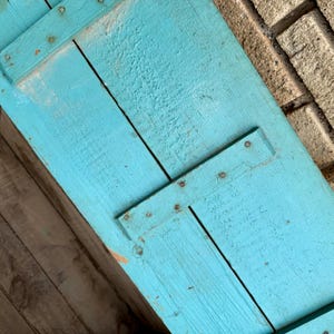 May include: Close-up of a section of a weathered, turquoise-painted wooden shutter. The shutter is constructed from planks held together with metal straps and rivets. The wood grain is visible, and the paint has a distressed appearance.