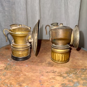 May include: Two antique brass miner's lamps. The lamps have a round body, a handle, and a large, curved reflector. The brass has a warm, aged patina. The lamps are displayed on a wooden surface.