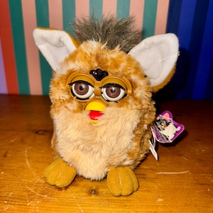 Könnte beinhalten: Ein Vintage-Furby-Spielzeug mit einem braunen und beigefarbenen Plüschäußeren, großen braunen Augen und einem gelben Schnabel. Das Spielzeug hat weiße Ohren und einen Büschel braunen Fells auf dem Kopf. Ein lila Etikett ist angebracht.