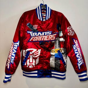 May include: A red satin Transformers bomber jacket with blue and white accents. The jacket features the Transformers logo and character illustrations, with the words "TRANSFORMERS" prominently displayed. The collar and cuffs have blue and white stripes.