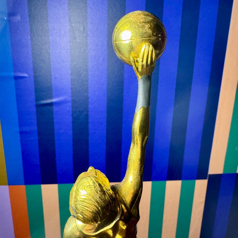 Puede incluir: Trofeo dorado que representa una figura sosteniendo una pelota por encima de la cabeza. La figura est&aacute; en una pose din&aacute;mica, con el brazo extendido hacia arriba. El trofeo tiene un acabado met&aacute;lico.