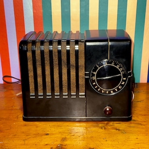 Puede incluir: Una radio Silvertone negra vintage con una rejilla de altavoz marrón y un dial redondo. La radio tiene una luz indicadora de encendido roja y un dial de sintonización con la palabra "Silvertone". La radio está sobre una superficie de madera.