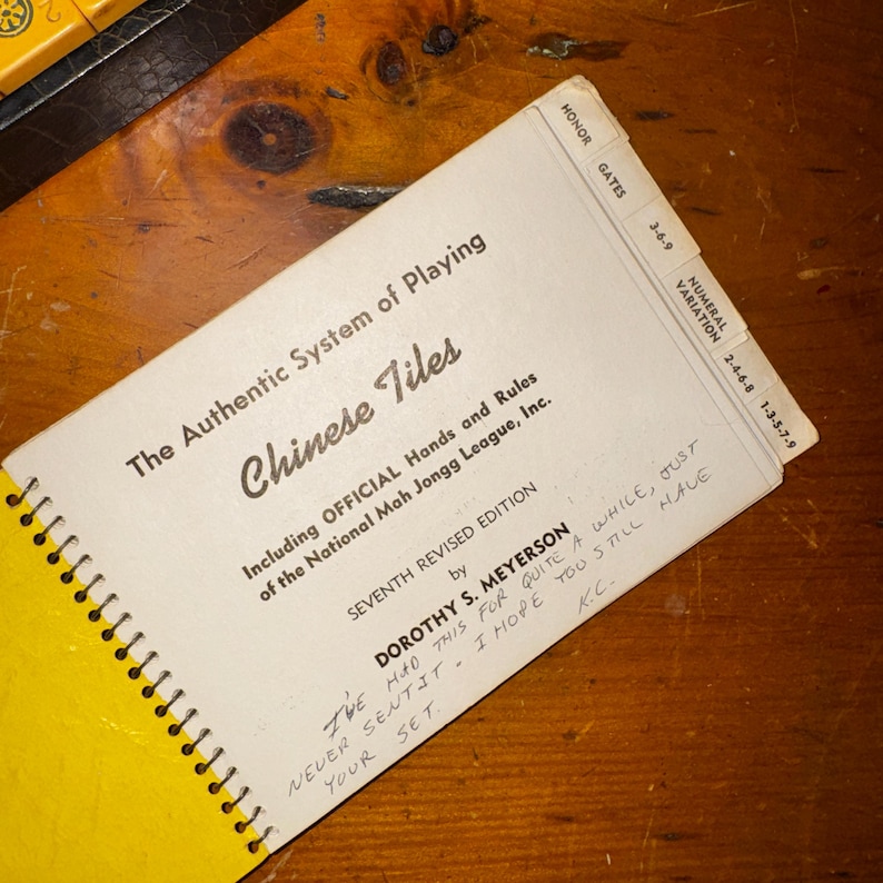 Op de afbeelding: Een spiraalgebonden boek met de titel "The Authentic System of Playing Chinese Tiles" met de tekst "Chinese Tiles" in een decoratief lettertype. Het boek bevat de tekst "Including OFFICIAL Hands and Rules of the National Mah Jongg League, Inc."